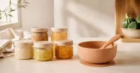 Illustration: plain sealed baby-food containers and an empty clean silicone bowl with spoon on a neutral kitchen surface, no infant, no feeding scene