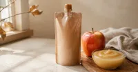 Illustration: unbranded sealed fruit puree pouch beside an apple and small glass bowl of smooth puree, no baby, no feeding scene