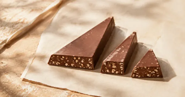 Illustration: three unbranded triangular chocolate bars of different lengths on cream paper, small nougat flecks visible Illustration: three unbranded triangular chocolate bars of different lengths on cream paper, small nougat flecks visible